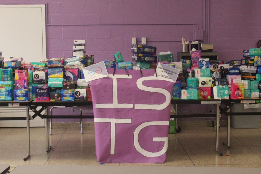 Donated menstrual products on top of a table with a sign in front saying, ISTG which stands for I Support the Girls